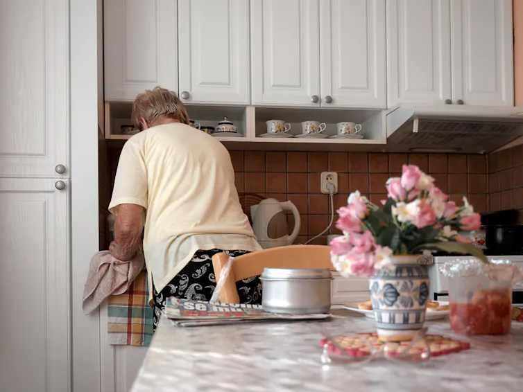 An older woman seen from behind in a light, airy kitchen.