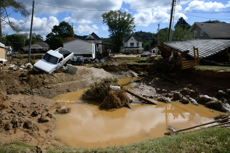 A big pool of muddy water surrounded by small homes and a white pickup truck that's angled into a ditch.