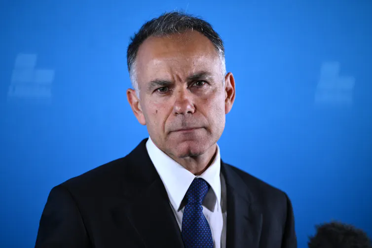 A man in a suit stands in front of a blue backdrop.