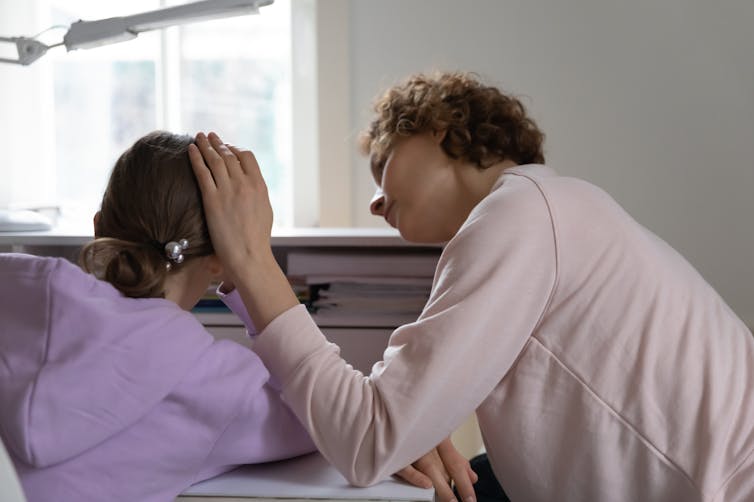 A woman comforts her teenage daughter, seen from behind.