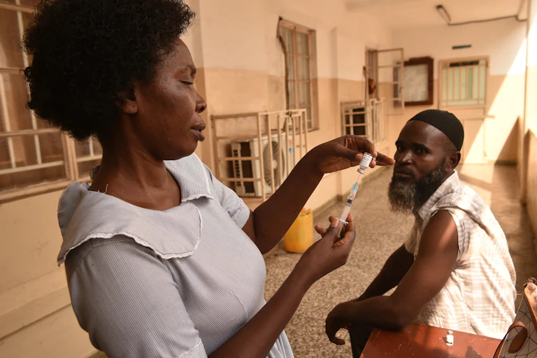 A woman fills a syringe from a small bottle while a man sits with his sleeve rolled up
