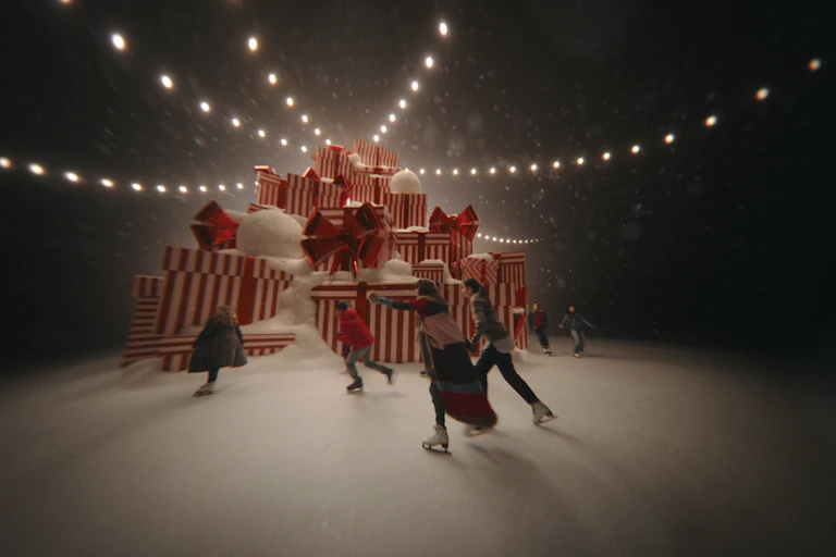 People ice skating around large pile of wrapped presents with fairy lights.