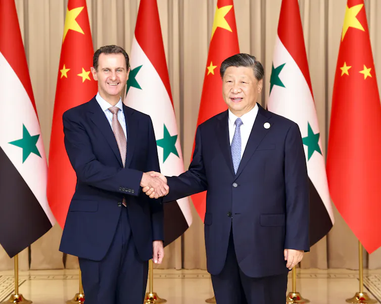 Bashar al-Assad shakes hands with Xi Jinping in front of their national flags.
