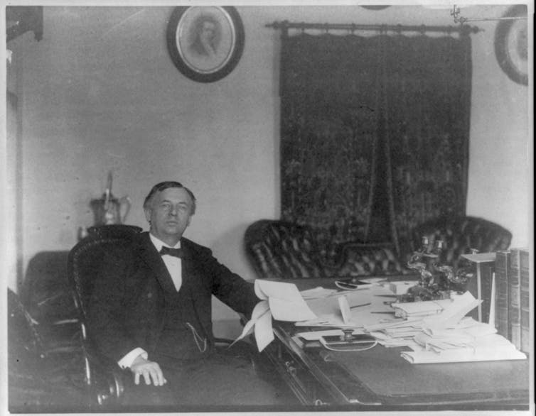 Picture circa 1890 of a man wearing a black suit, vest and bow tie sitting at large desk with leather sofa in background