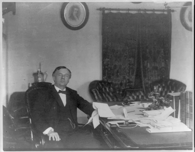 Picture circa 1890 of a man wearing a black suit, vest and bow tie sitting at large desk with leather sofa in background