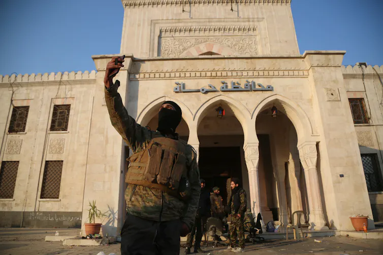 A Syrian rebel fighter takes a selfie in front of a government bulding while his comrades rest.