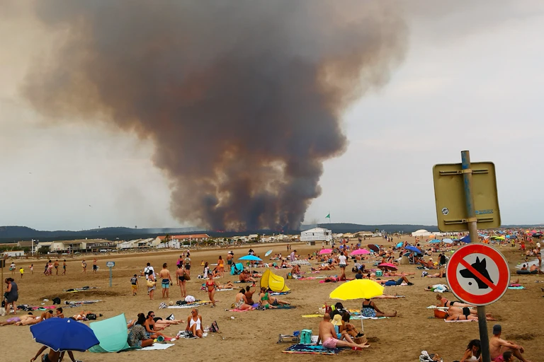 Plage où les touristes regardent une colonne de fumée noire géante provenant d'une forêt en feu près de Narbonne