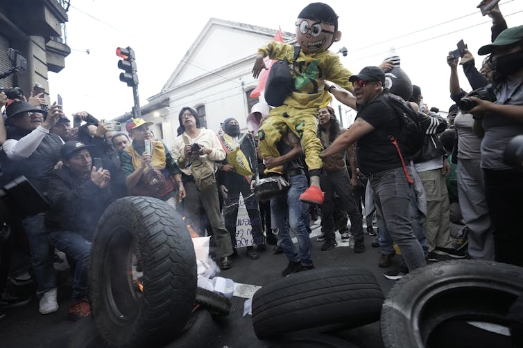 A man carries a human sized doll wearing yellow clothes and glasses, others around him take photos