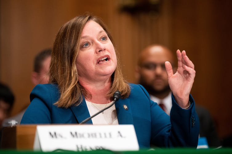 Middle-aged woman wearing blue suit jacket speaks in front of microphone