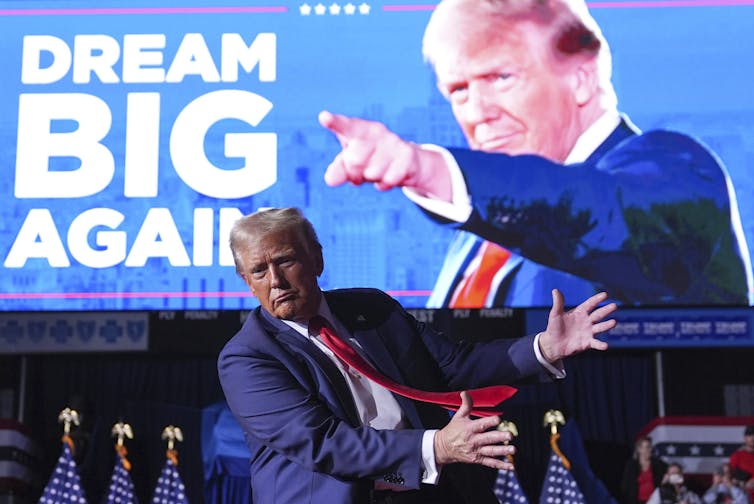 A man in a blue suit with greyish blond fluffy hair contorts his body in front of a sign that features a photo of him pointing and reads Dream Big Again.
