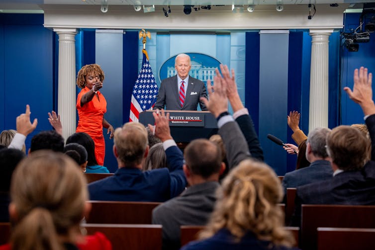 President Biden stands at the podium in the White House press room as reporters raise their hands