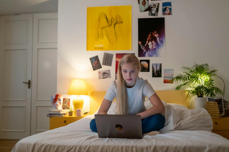 A teenage girl sits on a bed looking at a laptop.