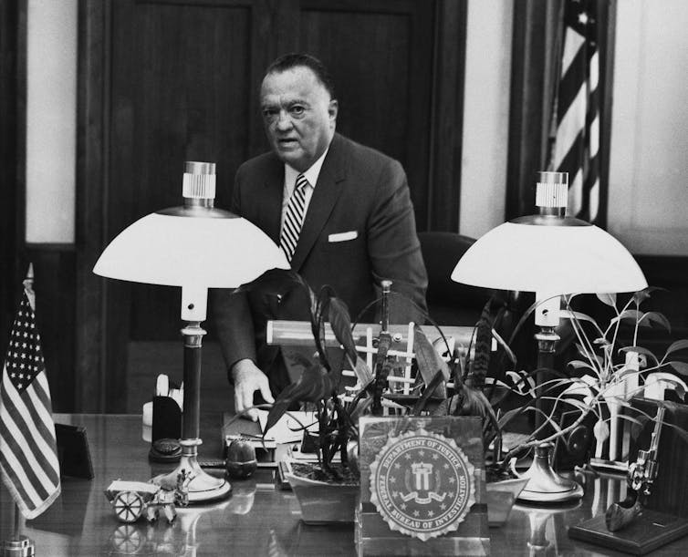 A man in a suit standing behind a desk covered with a lot of things.