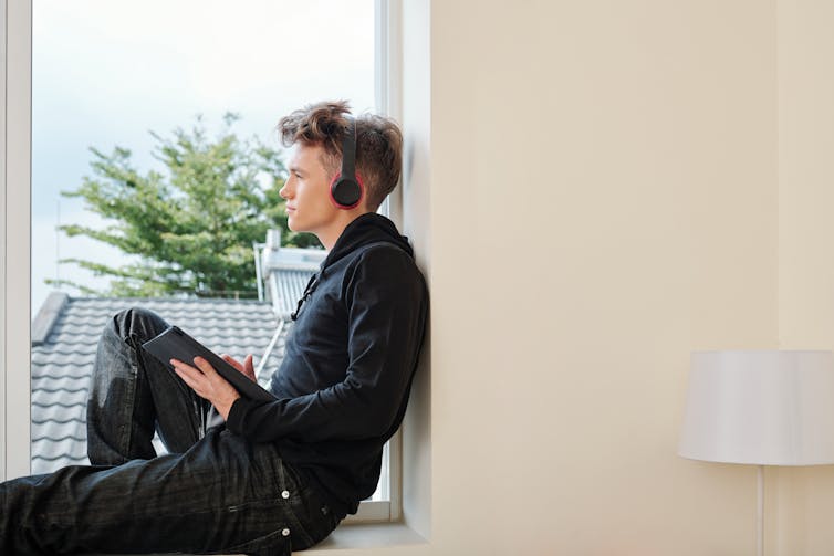 A young man wears ear phones and sits by a window.