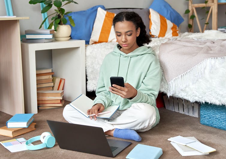 A teenager sits next to a bed, laptop and papers spread out in front of her. She holds a notebook, pen and phone.