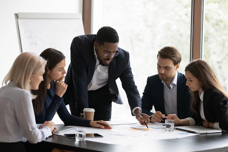 Confident boss points at documents on table surrounded by team on employees