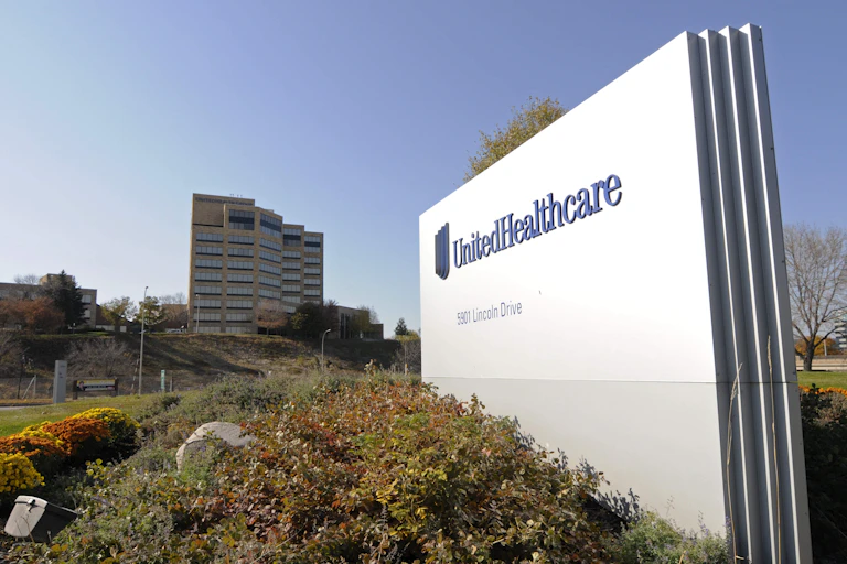 sign labeled unitedhealthcare sits in grass near flowers, in distance is a big building