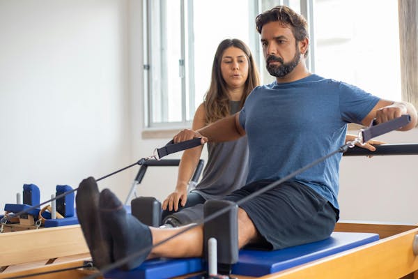 Man pulls straps of reformer, with his physio looking on