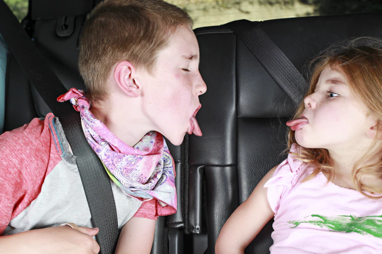 A brother and sister poke their tongues at each other in backseat of a car.