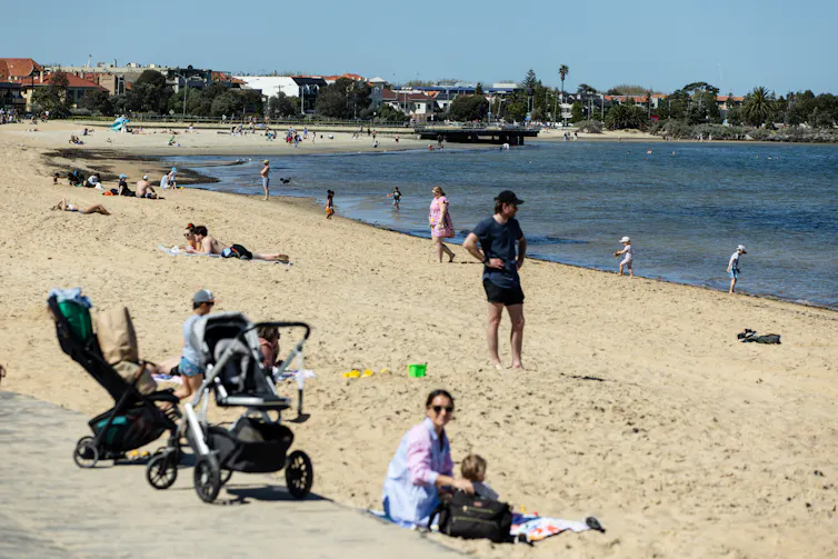 A mother and child sit near a pram at St Kilda beach in Melbourne. Small figures play near the water.