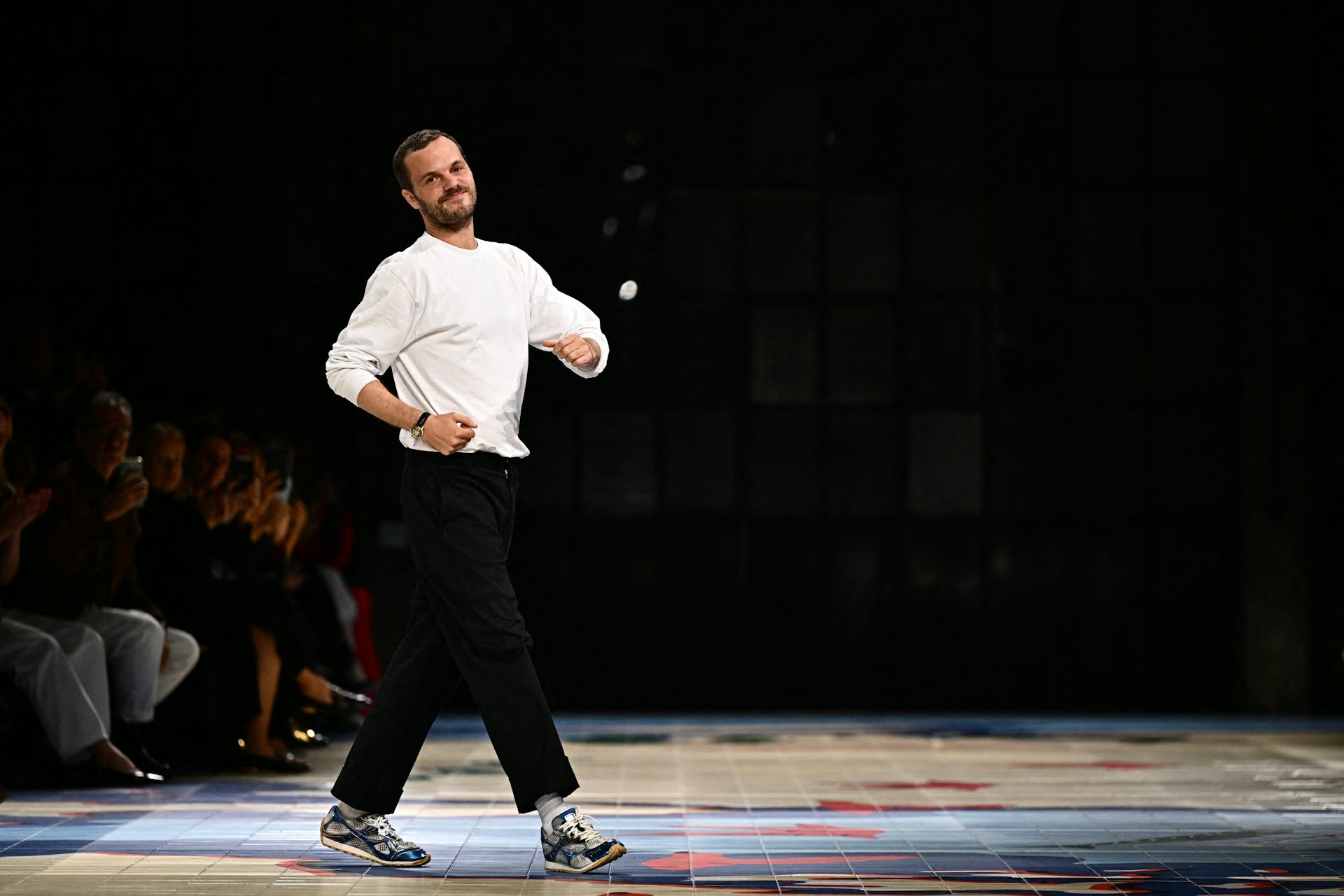 Mathieu Blazy at the end of a runway show, dressed in a white t-shirt and black pants, striking a playful pose.