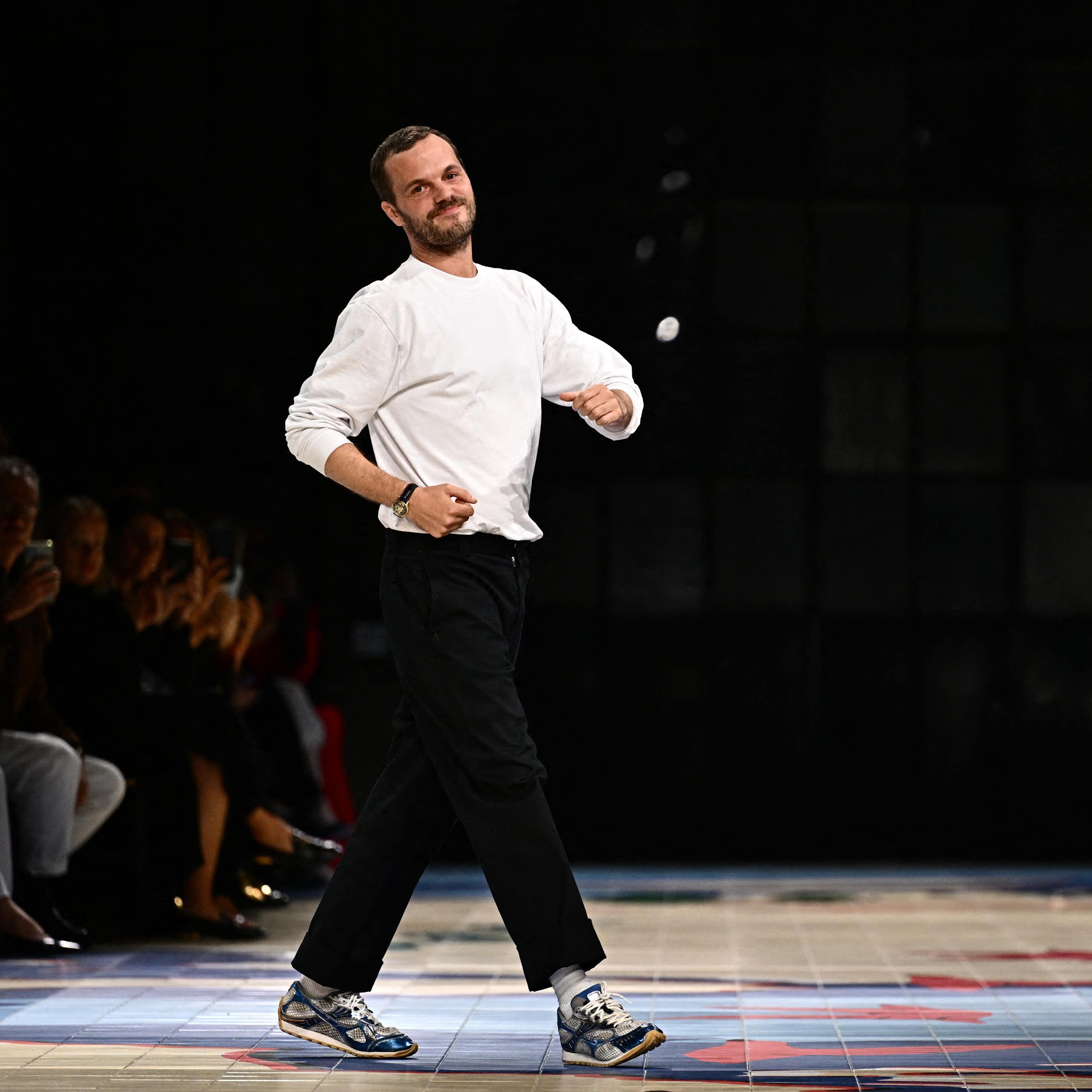Mathieu Blazy at the end of a runway show, dressed in a white t-shirt and black pants, striking a playful pose.