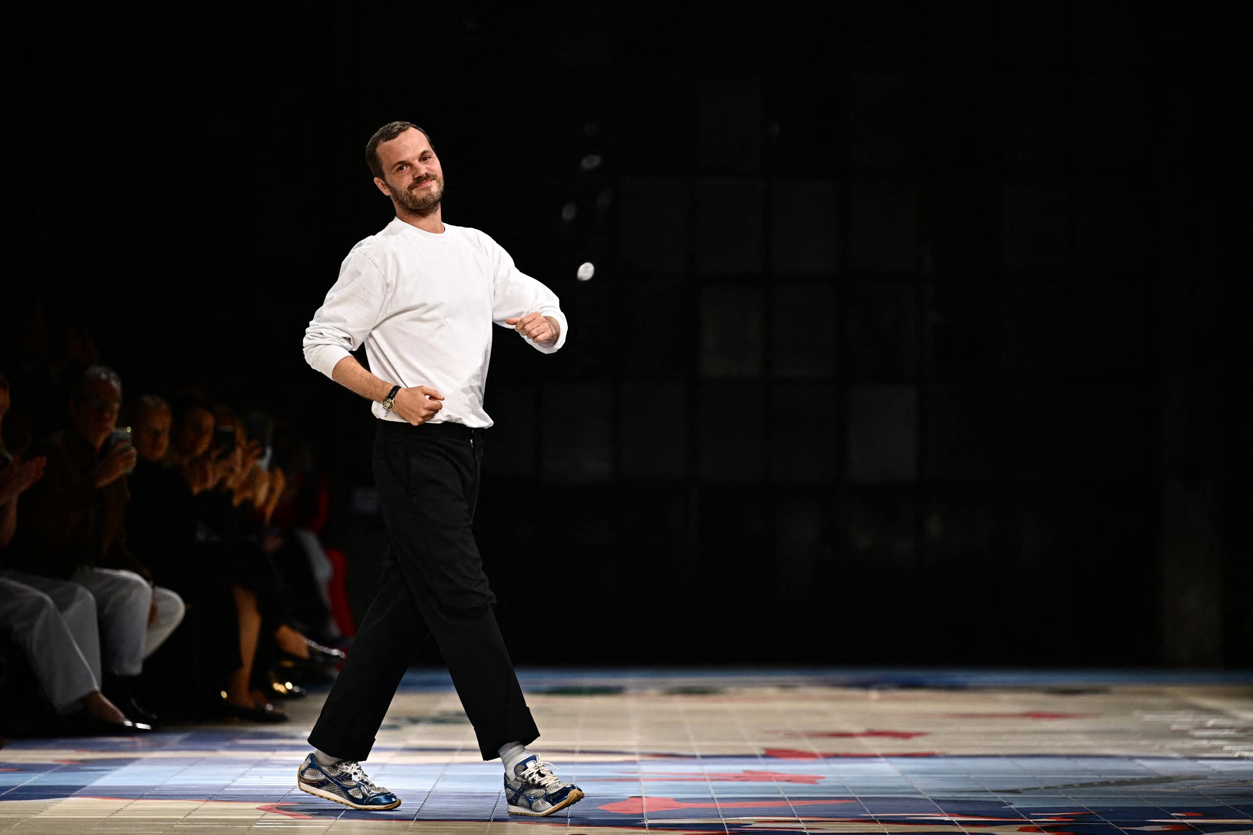 Mathieu Blazy at the end of a runway show, dressed in a white t-shirt and black pants, striking a playful pose.