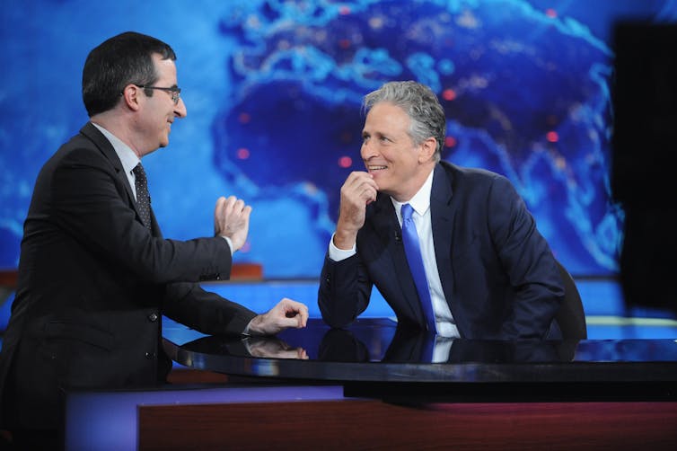 Two middle-aged men in suits sit at a news anchor's desk and share a laugh.