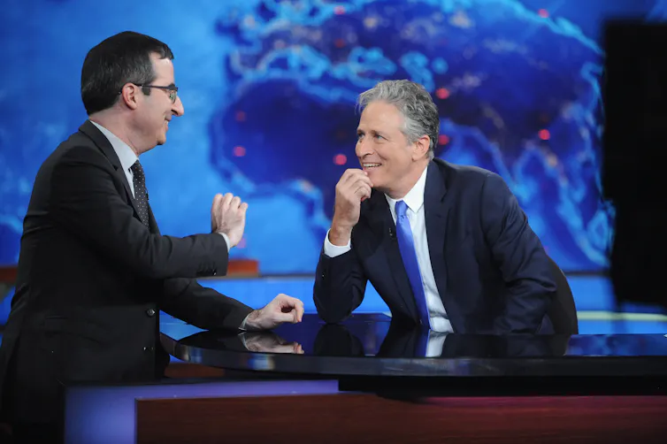 Two middle-aged men in suits sit at a news anchor's desk and share a laugh.
