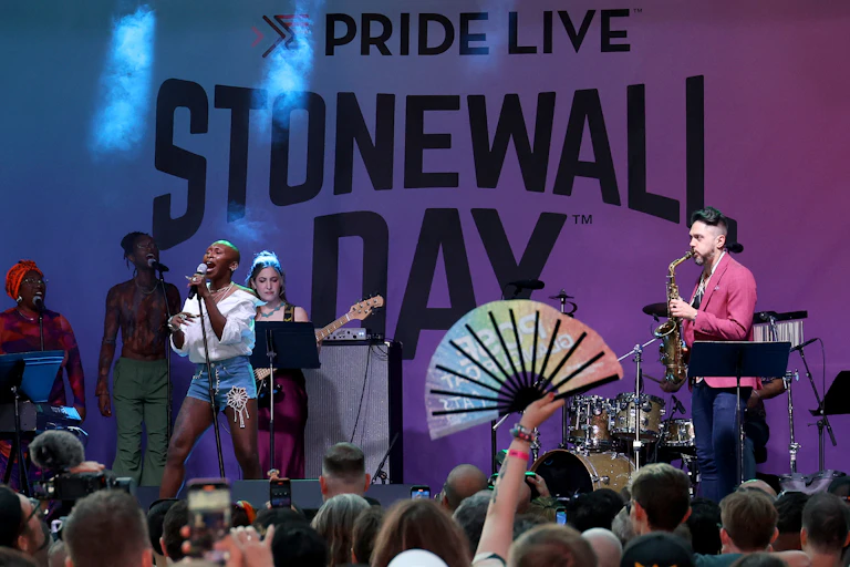 A woman with no hair sings in front of the words 'Pride Live Stonewall Day' before a big crowd.