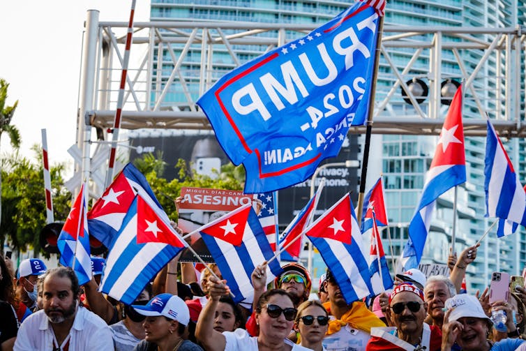 A Blue Trump flag flies above other flags and a crowd of people.