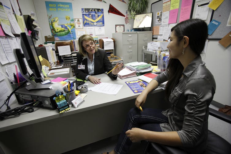 A woman behind a desk cluttered with lots of stuff talks to a female student