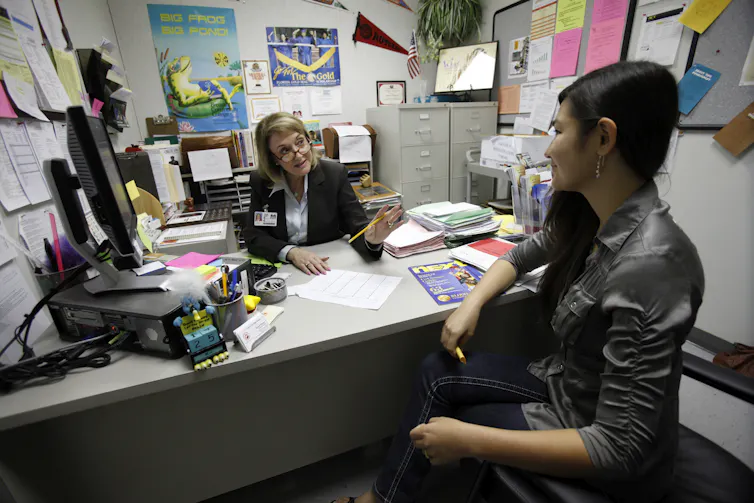 A woman behind a desk cluttered with lots of stuff talks to a female student