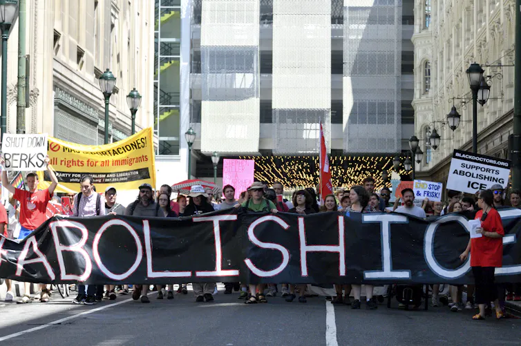 Crowd of people walk down a city street holding a large black banner that says 'Abolish ICE'