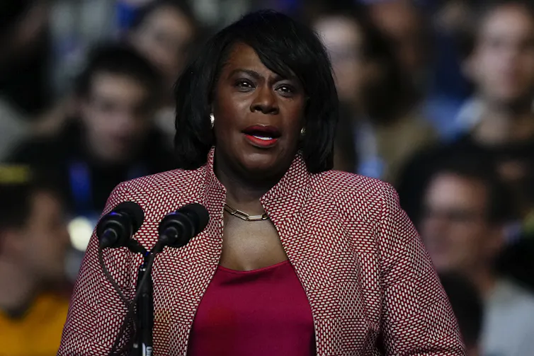 Woman in red shirt and red and white patterned jacket speaks in front of microphone to an audience