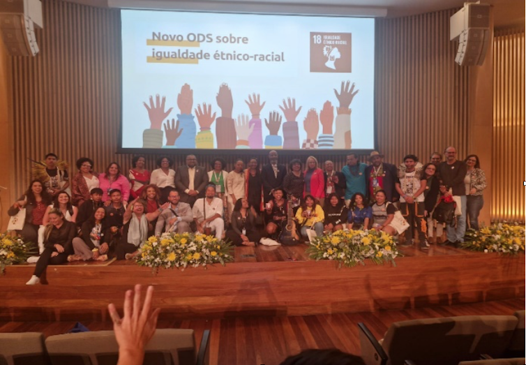 Várias pessoas posam para uma foto num auditório. Ao fundo lê-se numa tela grande Novo ODS sobre igualdade racial, com a ilustração de várias mãos, de raças diferentes, levantadas
