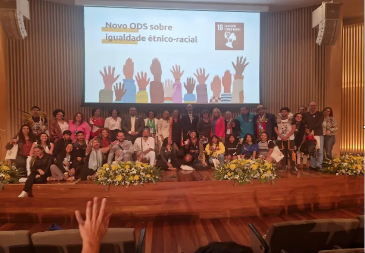 Várias pessoas posam para uma foto num auditório. Ao fundo lê-se numa tela grande Novo ODS sobre igualdade racial, com a ilustração de várias mãos, de raças diferentes, levantadas