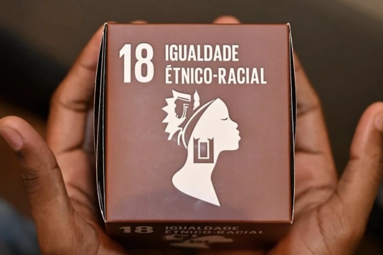 Mãos negras seguram um cubo marrom onde se lê: 18 Igualdade étnico-racial e o ícone do perfil de uma mulher com cabelos e brincos que trazem referências étnicas negras e indígenas