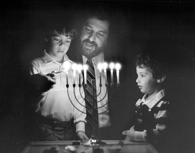 A black and white photo of a man in a suit and tie holding helping two young children light a candelabra with a flame.