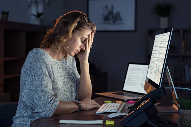 A woman sitting in front of a computer at a desk rests her head against her hand