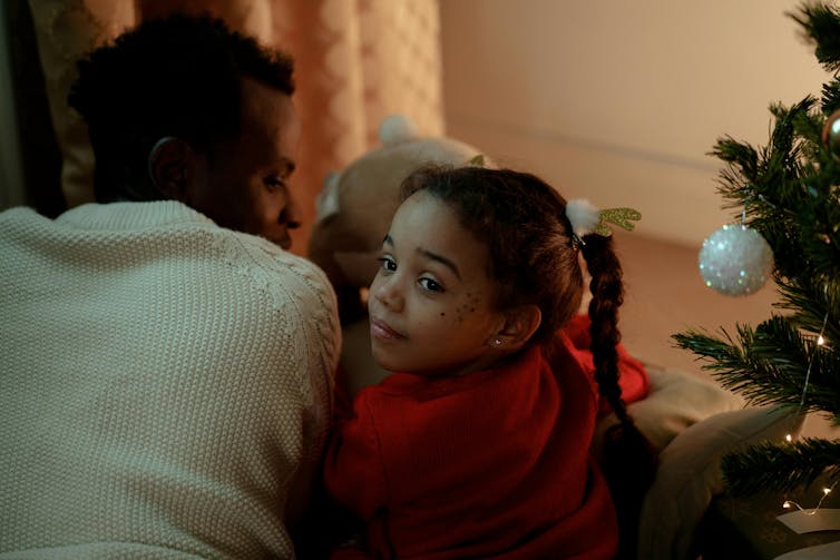 A young girl sits next to a man, next to a Christmas tree.