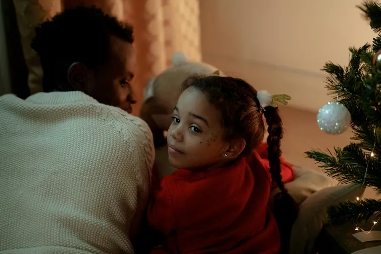 A young girl sits next to a man, next to a Christmas tree.