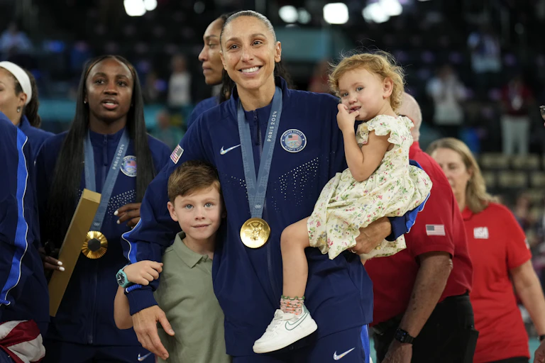 A woman wearing a gold medal around her neck carries a young girl with a young boy standing beside her
