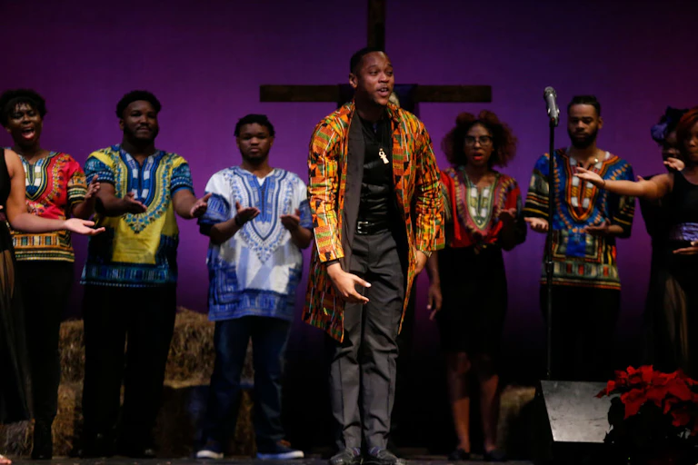 Half a dozen performers in brightly patterned tunics stand on a stage with a bright purple backdrop and a cross in the background.