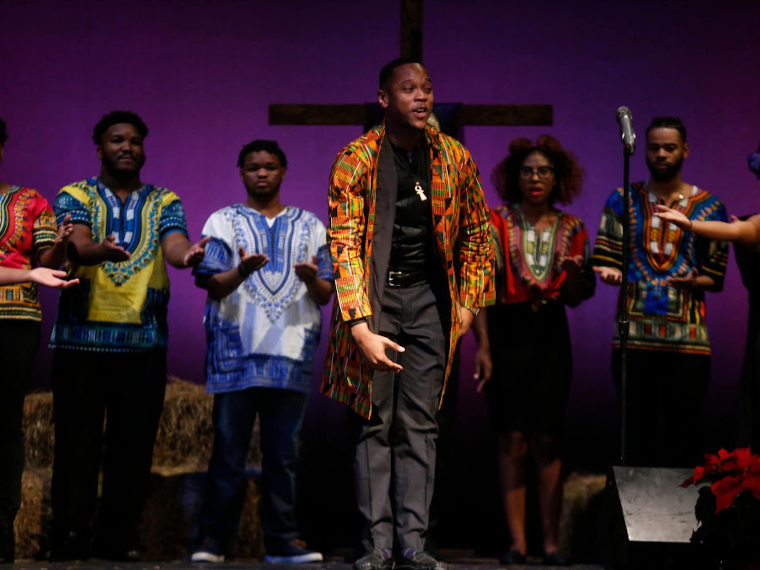 Half a dozen performers in brightly patterned tunics stand on a stage with a bright purple backdrop and a cross in the background.