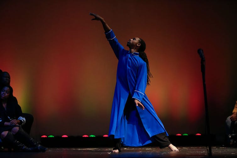 A man in a long, bright blue tunic poses elegantly on stage against an orange backdrop.