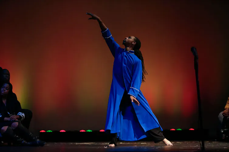 A man in a long, bright blue tunic poses elegantly on stage against an orange backdrop.