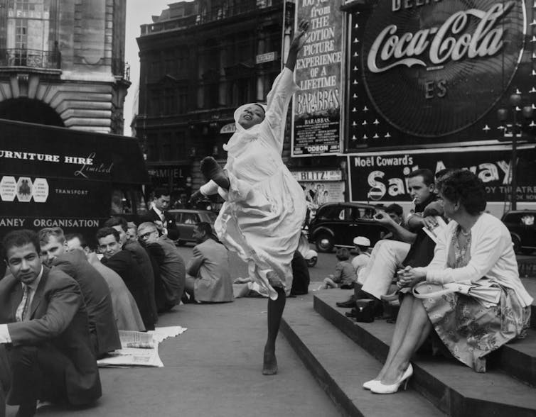 A woman in a white dress and black stockings dances among a crowd of people sitting outside in a city plaza.