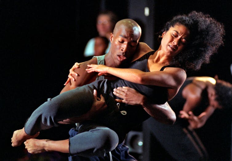 A man in a tank top lifts a woman in black pants and a black tank top as they rehearse a dance.