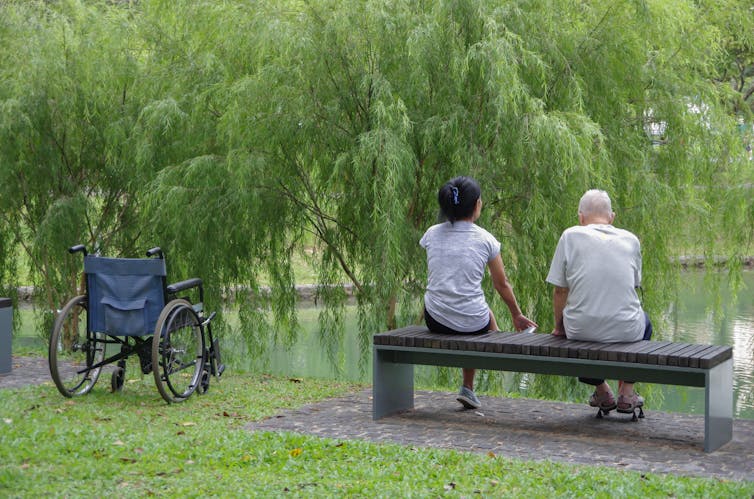 Two people sitting on a bench in a park overlooking a lake. An empty wheelchair is next to them.
